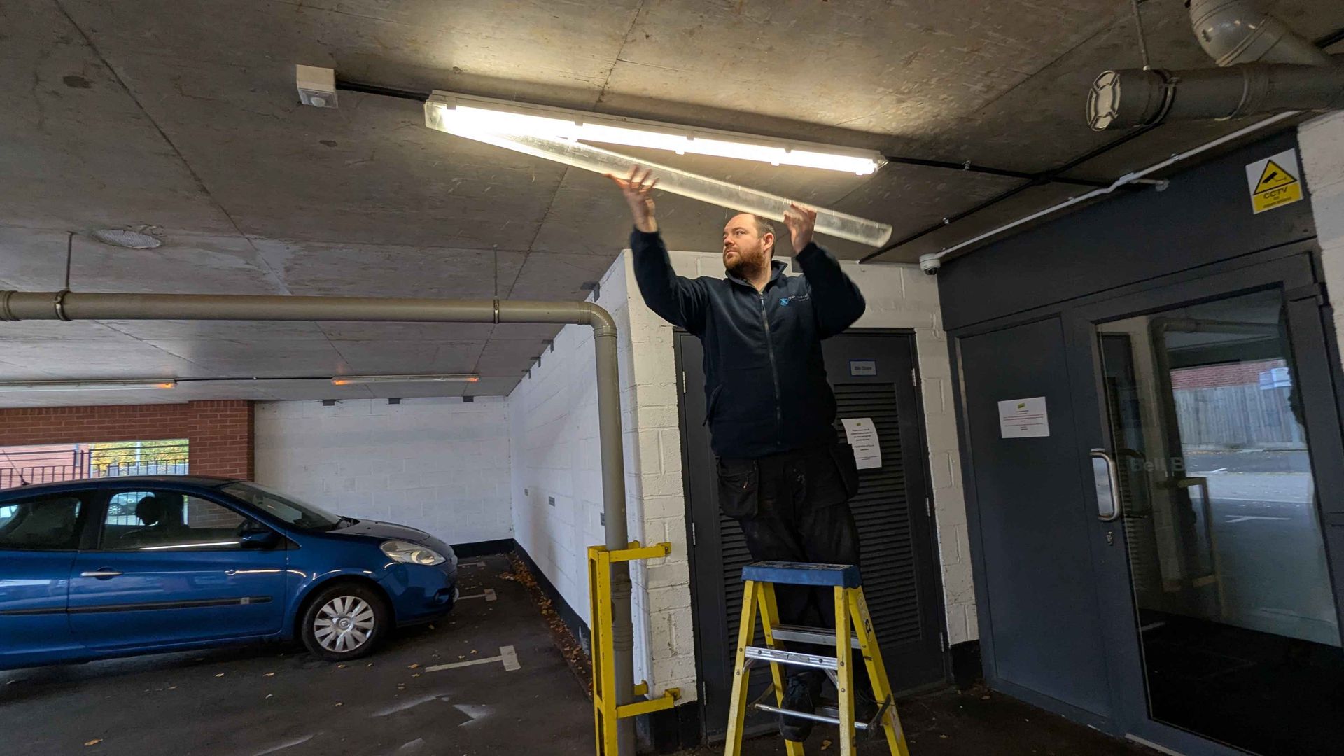 Man on a stepladder installing a fluorescent light fixture in a parking garage.