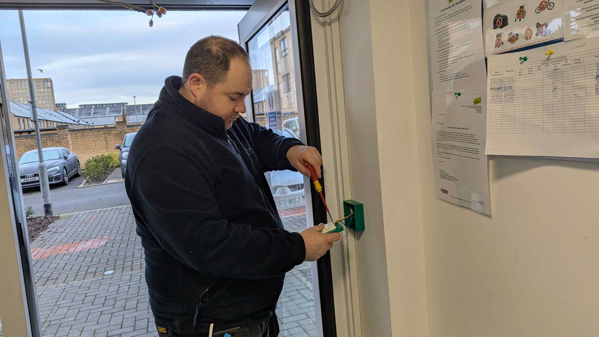 Man in black jacket uses a screwdriver on a green electrical box next to a door.