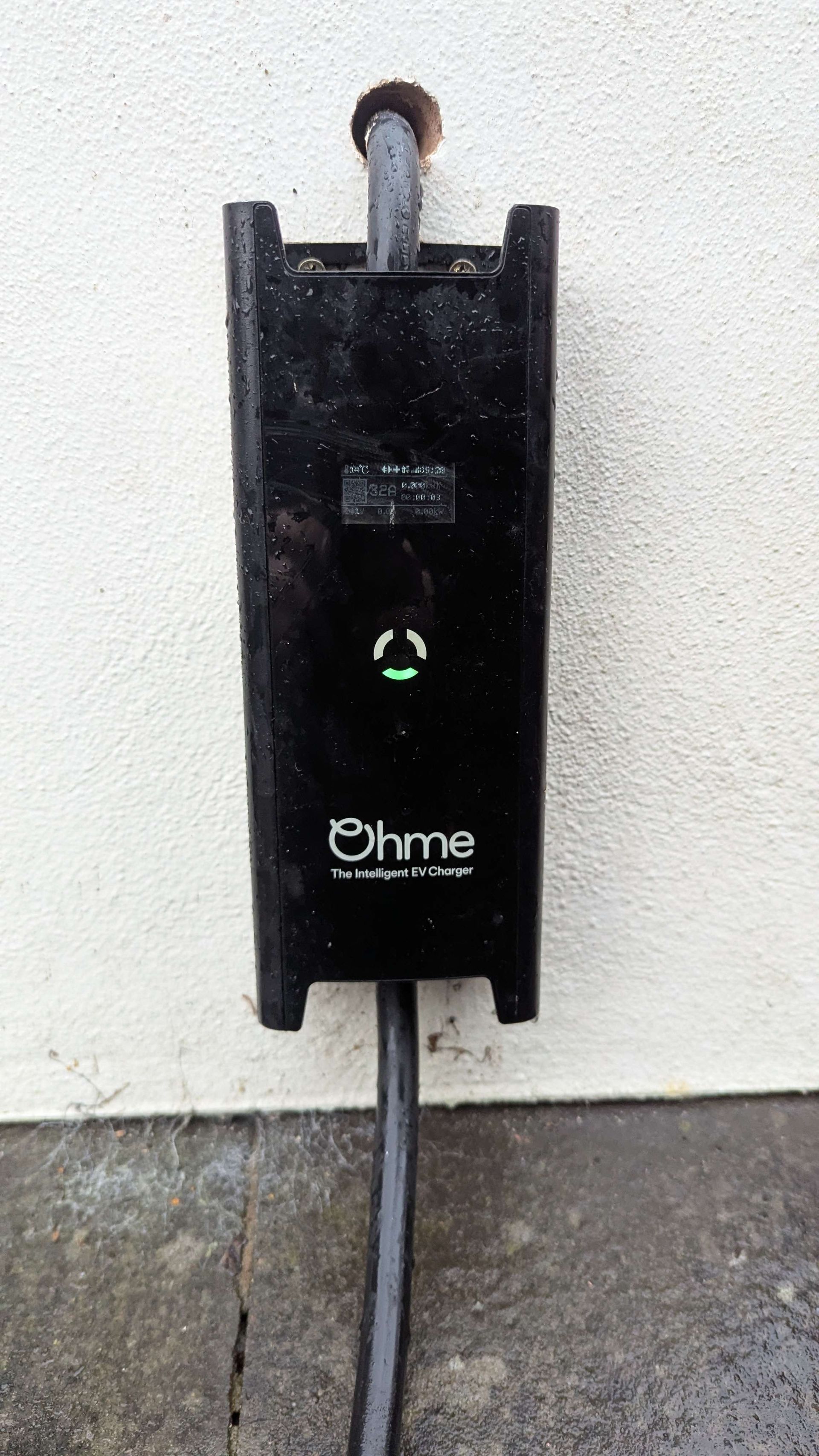 Black electric vehicle charger mounted on a white wall, with a green light illuminated.