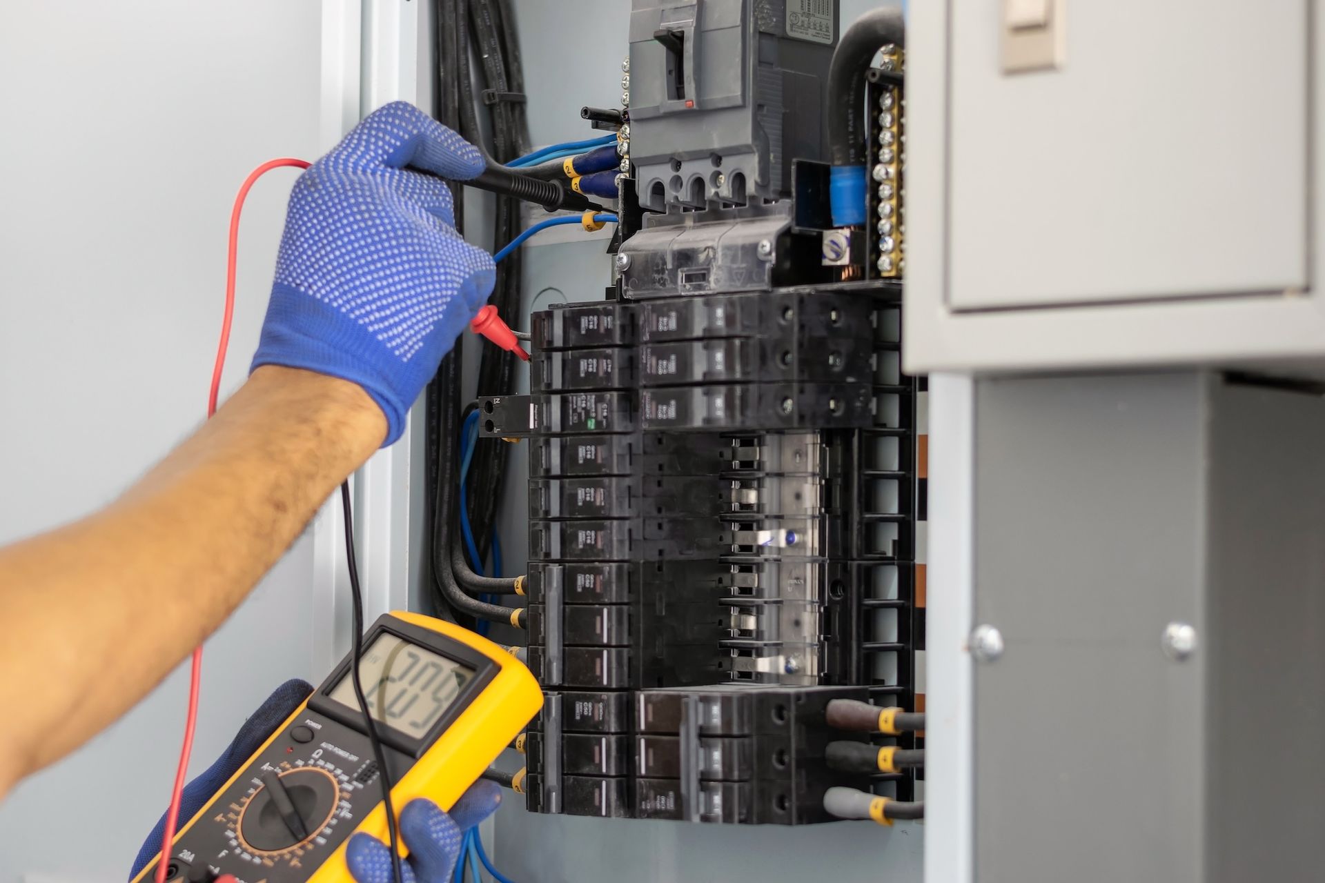 Electrician in blue gloves tests a circuit breaker panel with a multimeter.