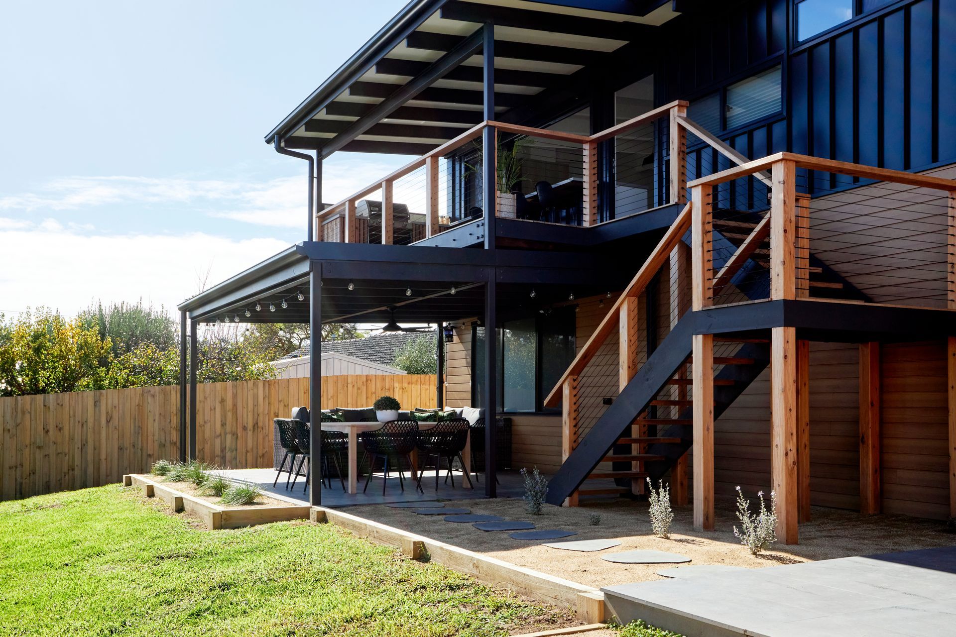 Two-story deck with dining area, stairs, and glass railing on a modern home exterior, surrounded by green grass and a fence.