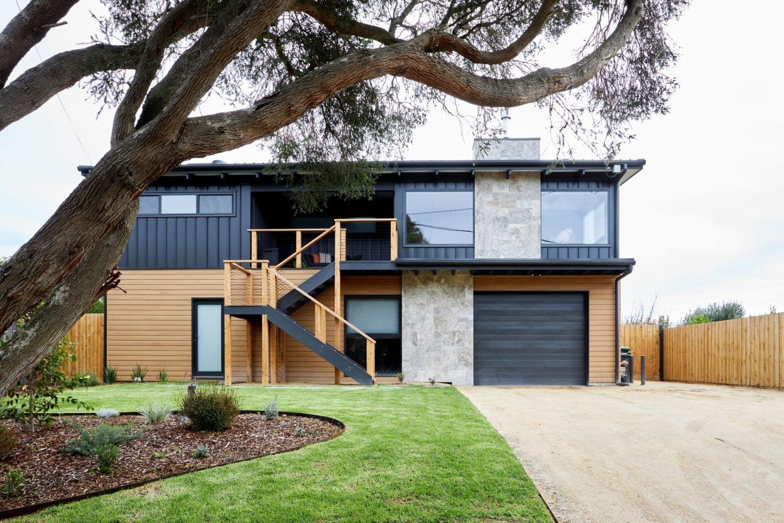 Modern two-story house with black and tan siding, stone accents, and a garage, under a tree.