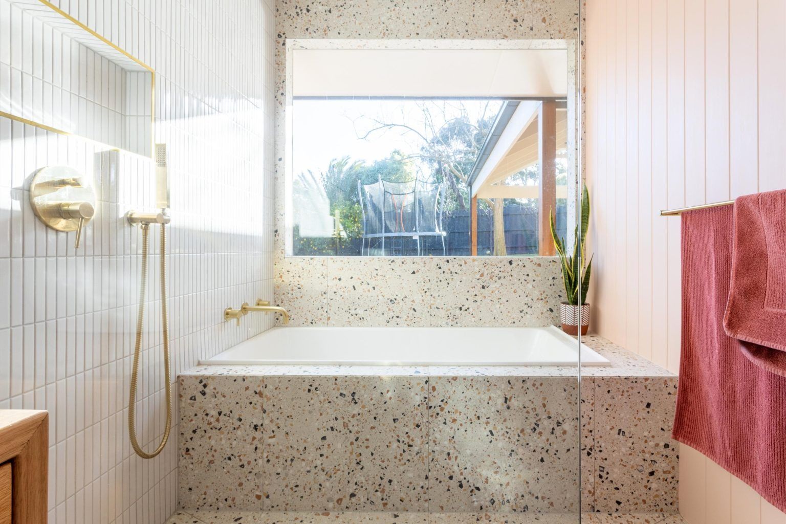 Modern bathroom with a bathtub, shower, and window overlooking greenery. Beige terrazzo, white tile, and gold fixtures.