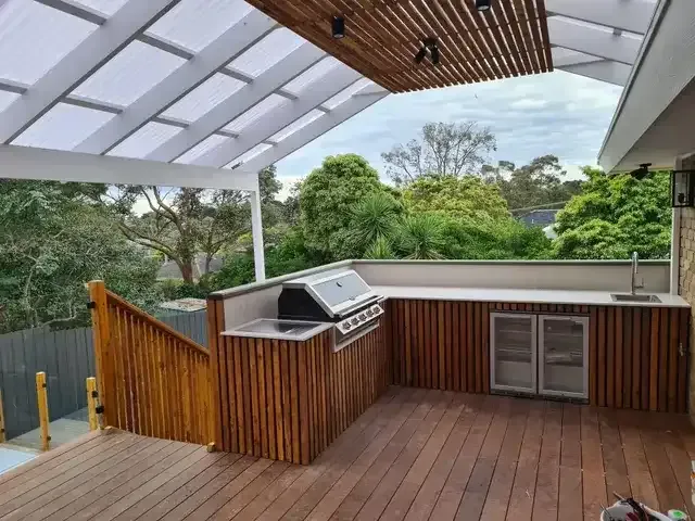 Outdoor kitchen with wooden cabinets, grill, sink, and mini fridge on a deck, with trees in the background.
