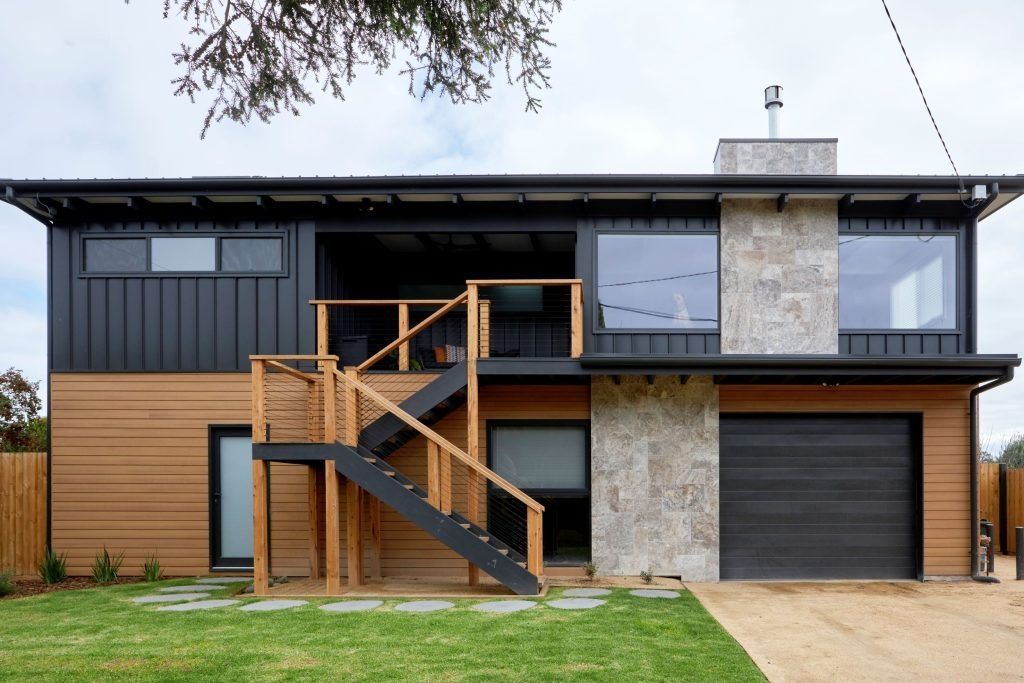 Two-story modern home with wood and black siding, stairwell, and garage. Green lawn and gravel driveway.