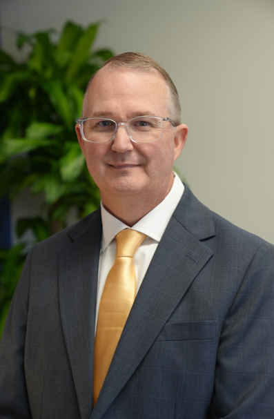 Man in a gray suit and glasses, smiling, outdoors in front of trees.