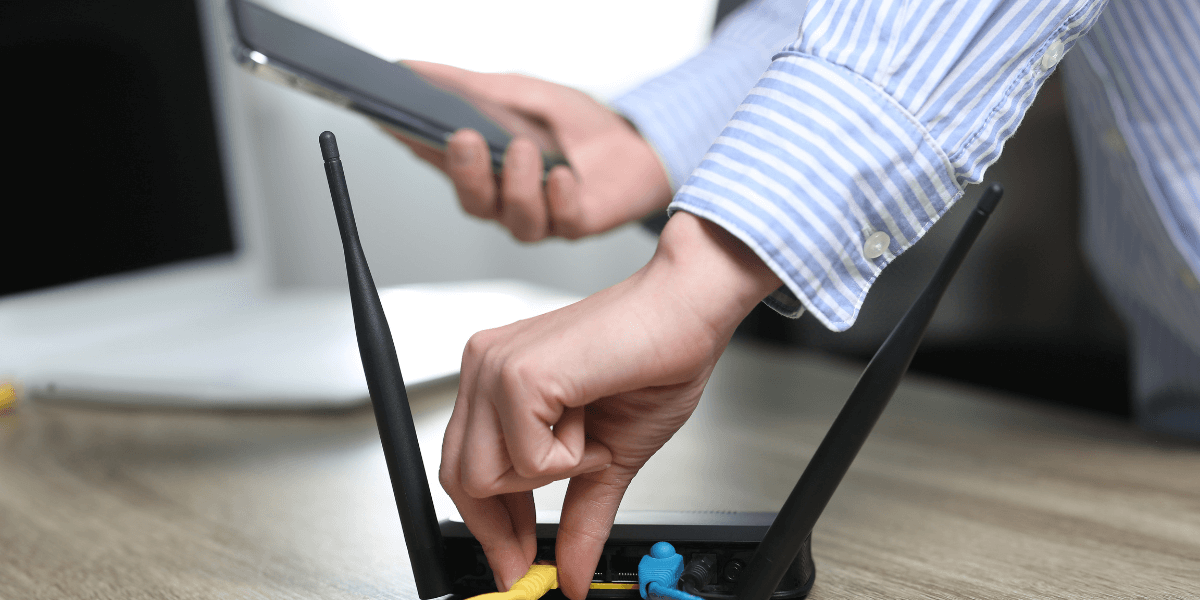 Hands connecting cables on a black Wi‑Fi router on a wooden table, with a laptop in the background