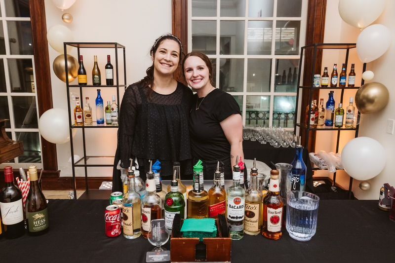 Two Women Are Standing in Front of A Bar Filled with Bottles of Alcohol - Lee’s Summit, MO - Top Shelf Bartending Services, LLC
