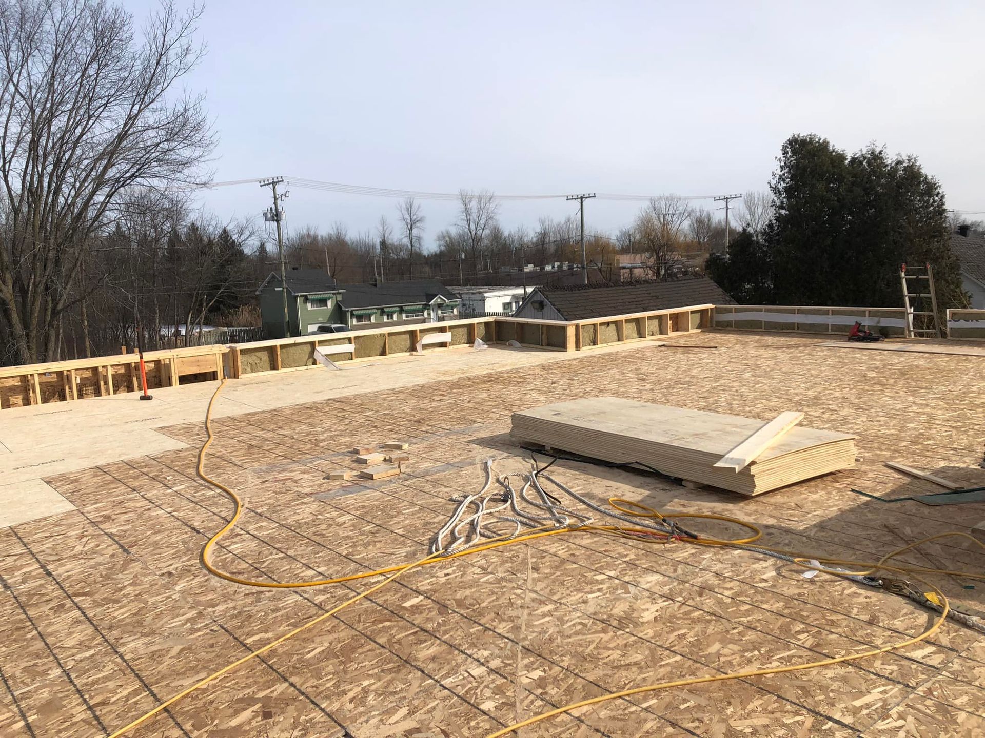 Chantier de construction avec sous-plancher en bois, murs partiellement ossature et un tas de matériaux de construction sous un ciel dégagé.