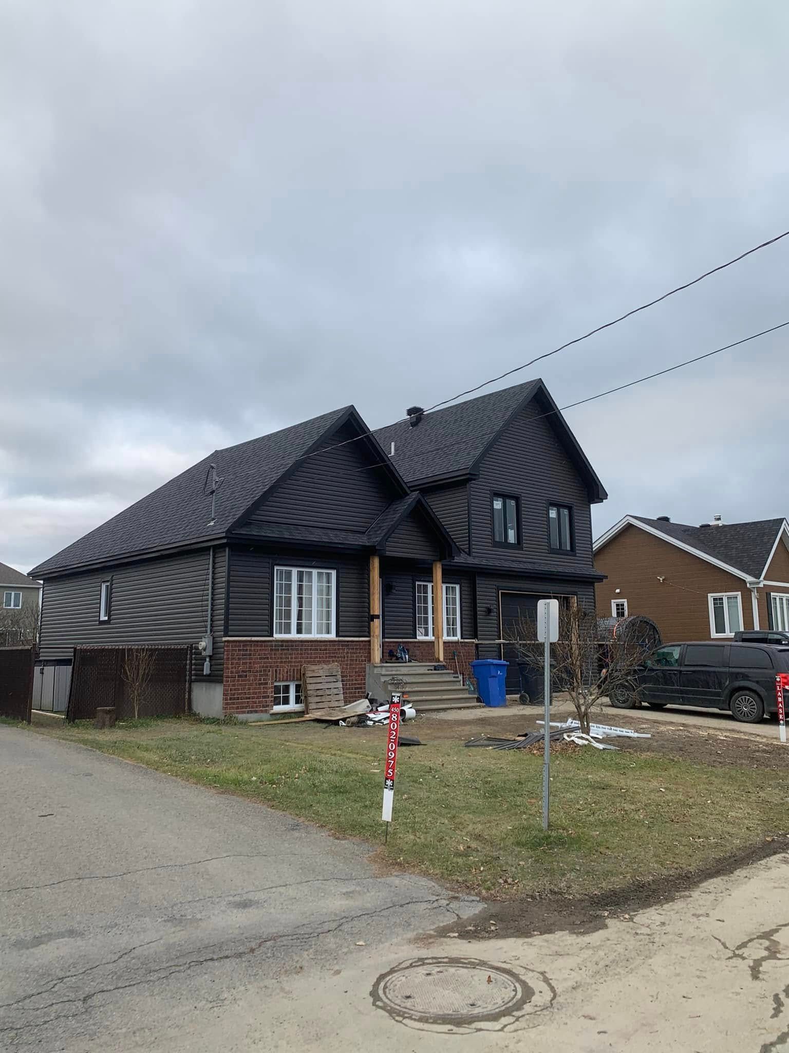Une maison de banlieue à deux étages avec un toit et un bardage en métal noir, des fondations en briques et une cour avant gazonnée.