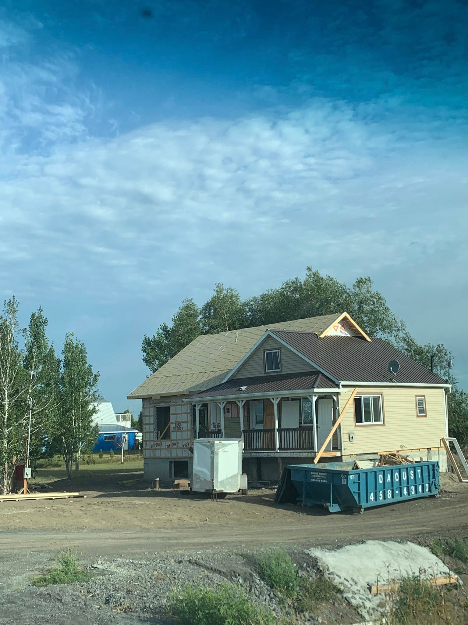 Une maison en rénovation avec une benne à ordures bleue devant, se détachant sur un ciel lumineux et nuageux et entourée d'arbres.