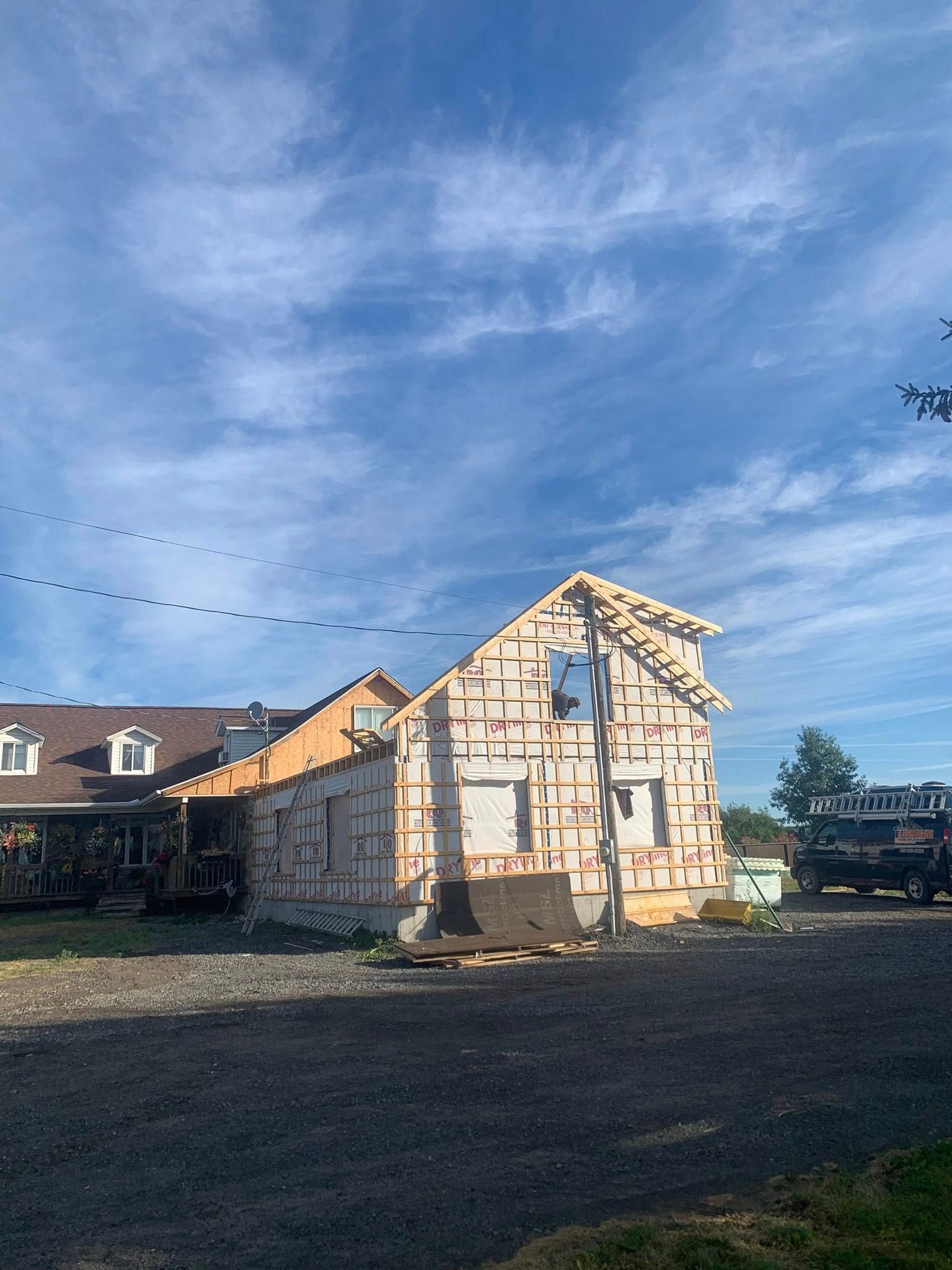 Une structure en bois à moitié construite se détache sur un ciel bleu, la partie achevée de la maison étant visible à gauche.