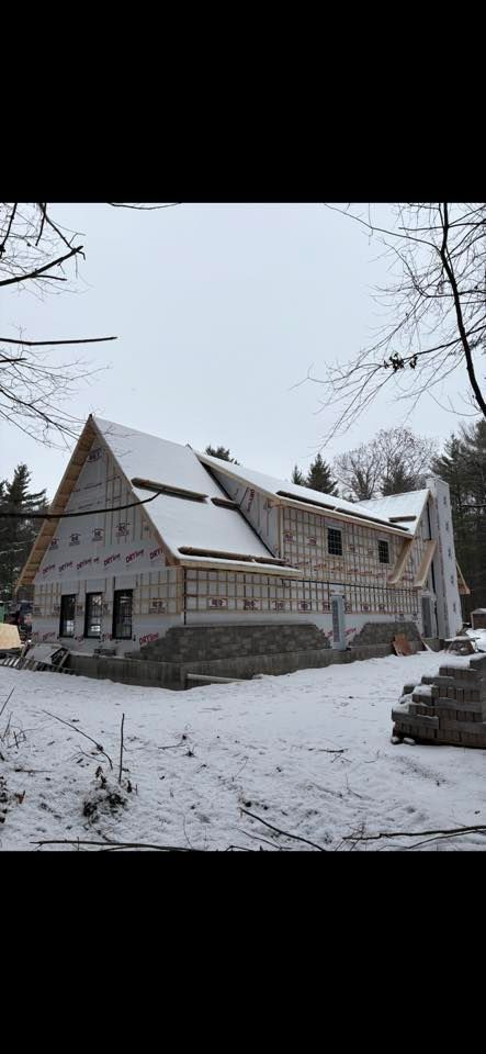 Une maison en construction, avec des fondations en pierre, une charpente en bois et un toit enneigé, dans un cadre boisé et enneigé.