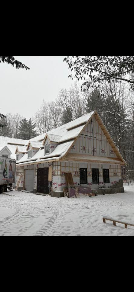 Une maison en construction, avec un revêtement apparent et un toit enneigé, située dans un environnement boisé et enneigé.
