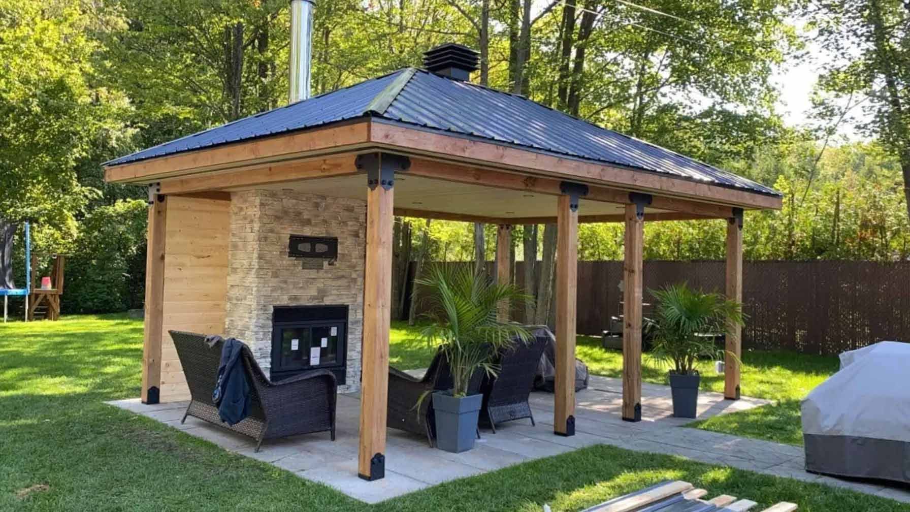 Pavillon extérieur avec cheminée en pierre, poteaux en bois et toit sombre, installé sur une terrasse en pierre dans un jardin gazonné.