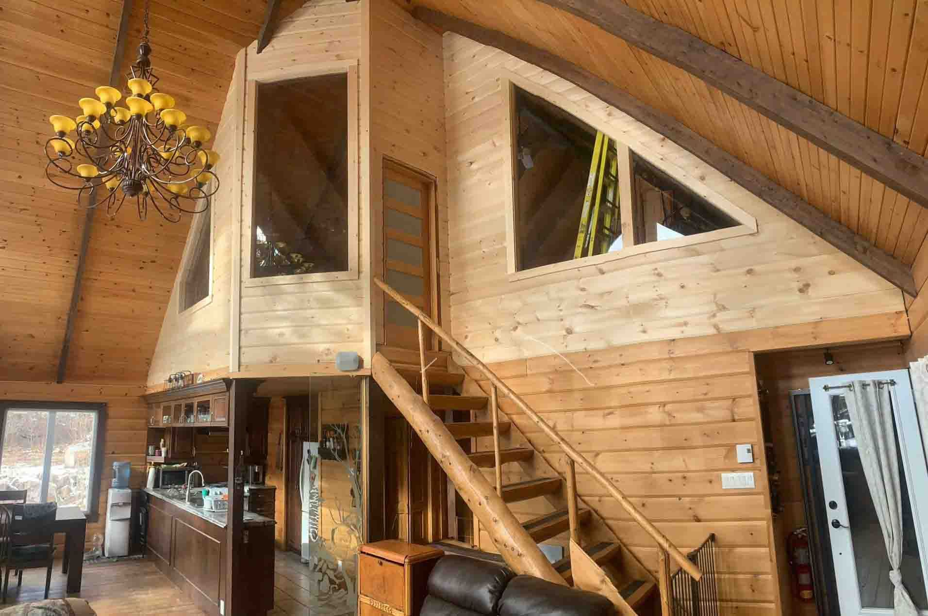 Intérieur d'une cabane en rondins rustique avec un escalier en bois, de hauts plafonds, de grandes fenêtres et un lustre décoratif.