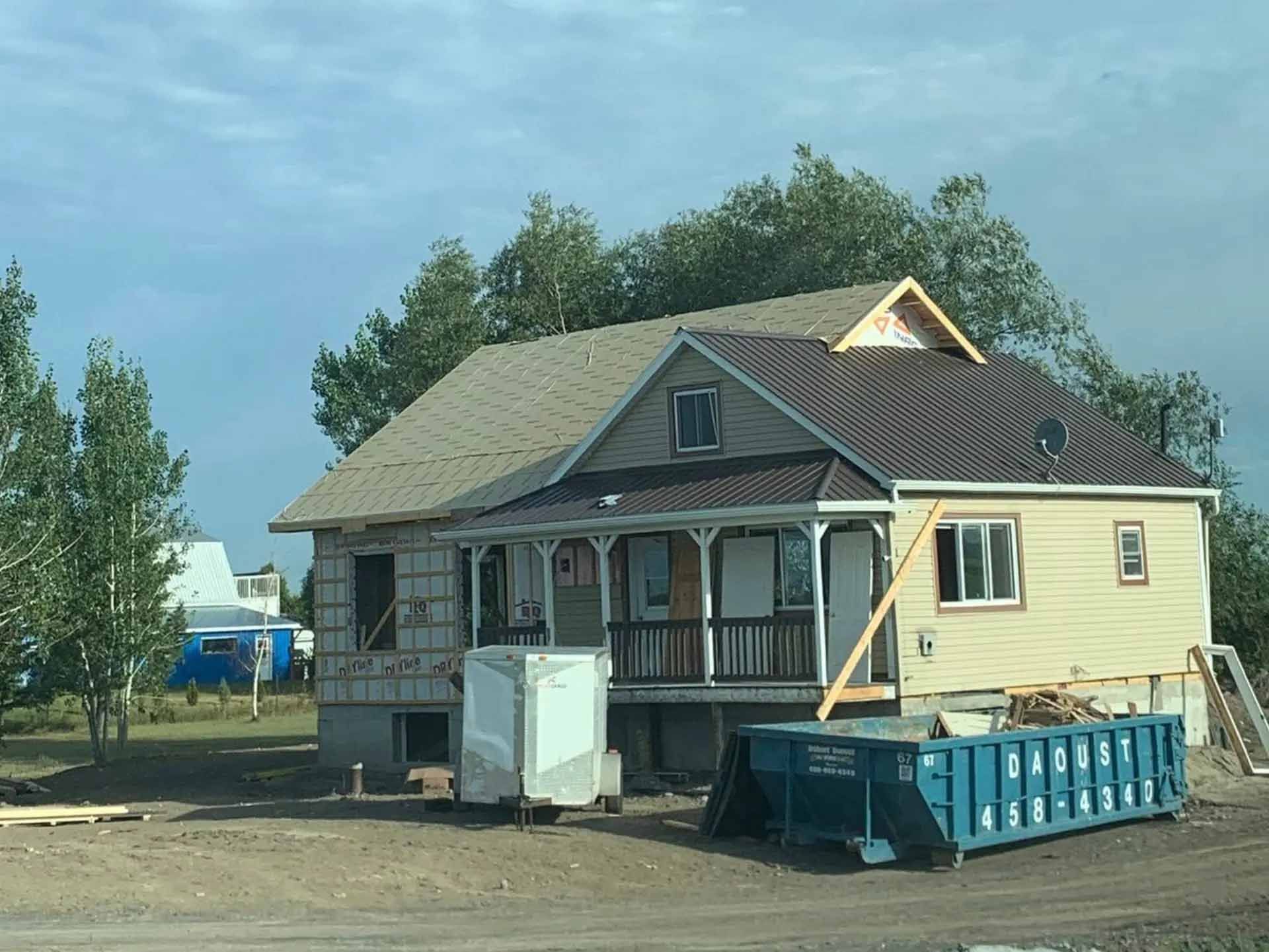 Une maison en rénovation avec un revêtement extérieur inachevé, un porche, une remorque de stockage portable et un conteneur à déchets bleu dans la cour.