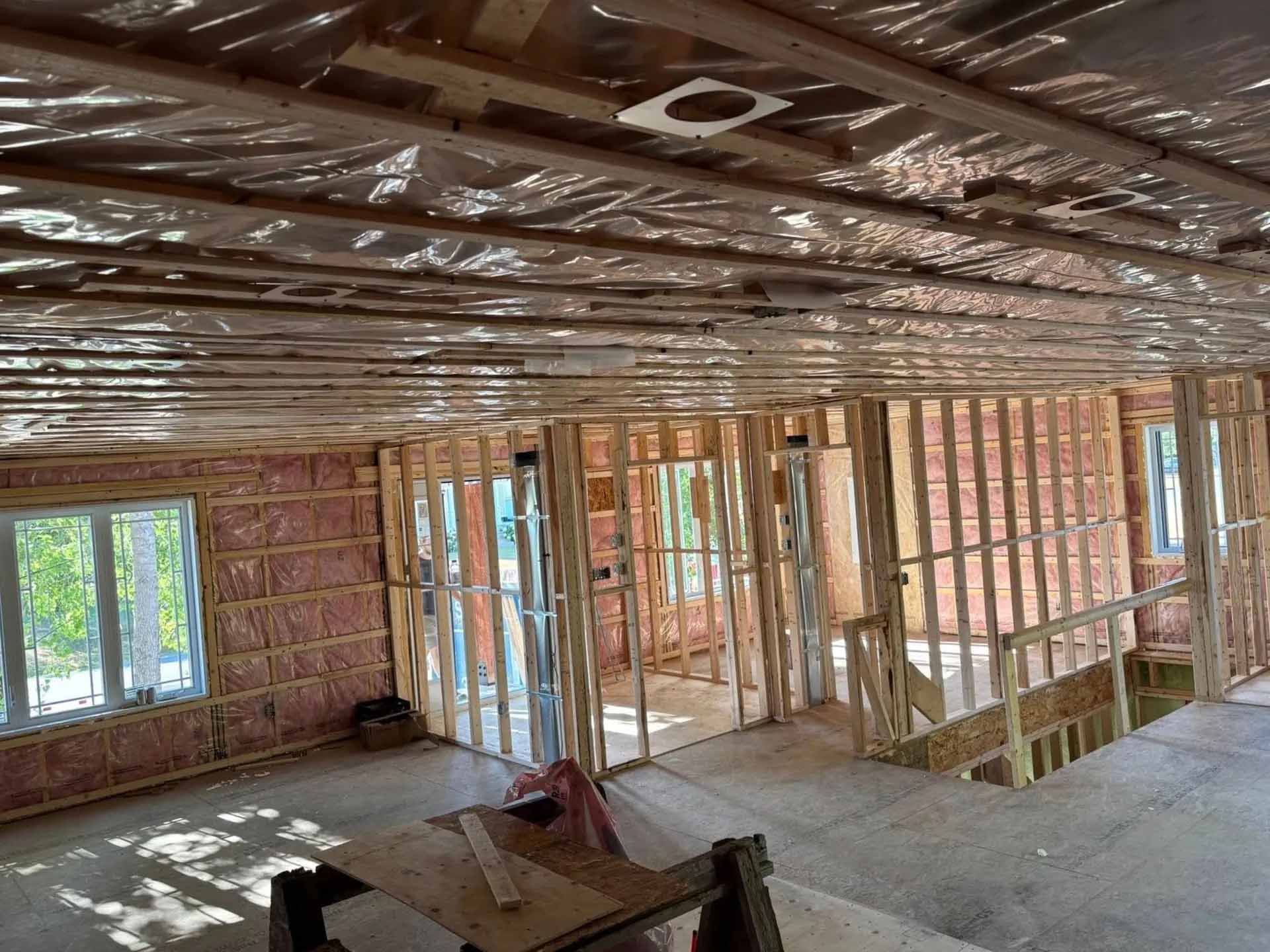 Vue intérieure d'un chantier de construction résidentielle avec des montants en bois apparents, une isolation rose et un plafond réfléchissant.