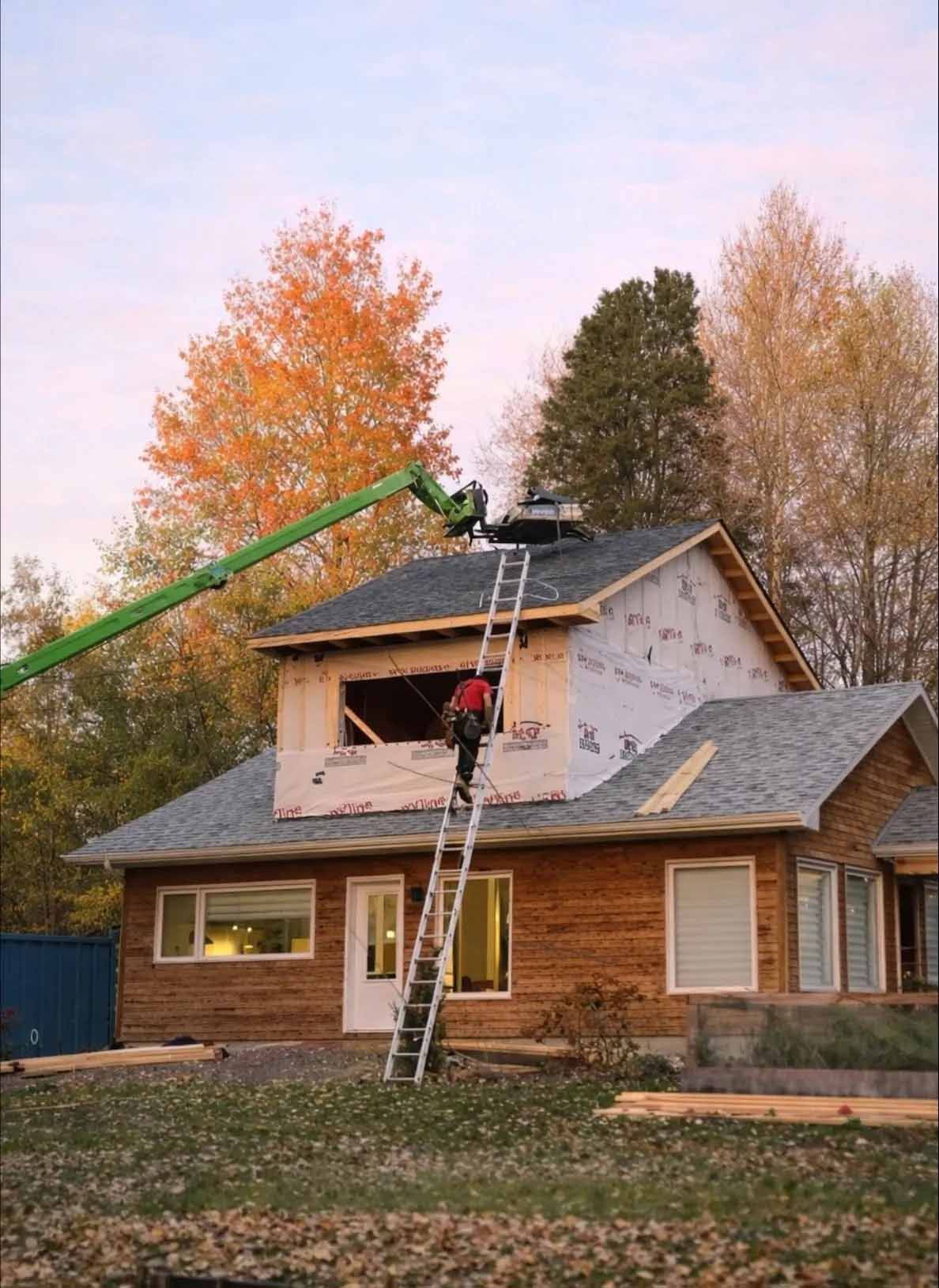 Un ouvrier grimpe à une échelle vers un toit en construction, aidé par le bras d'une grande grue verte, sur fond d'arbres d'automne.