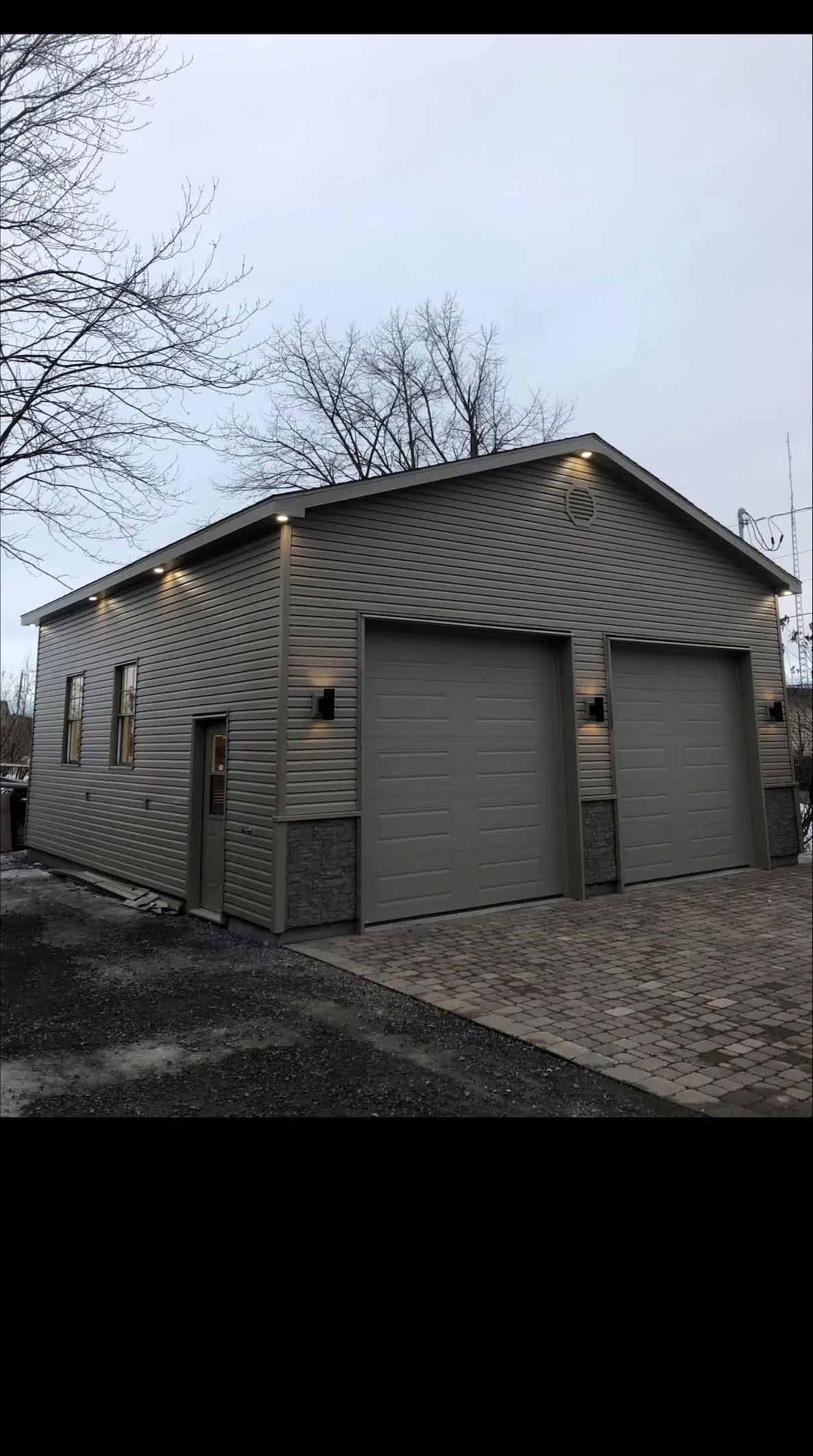 Un garage indépendant, revêtu de bardage gris et agrémenté de pierres, est doté de deux portes de garage et d'un éclairage extérieur qui s'allume au crépuscule.