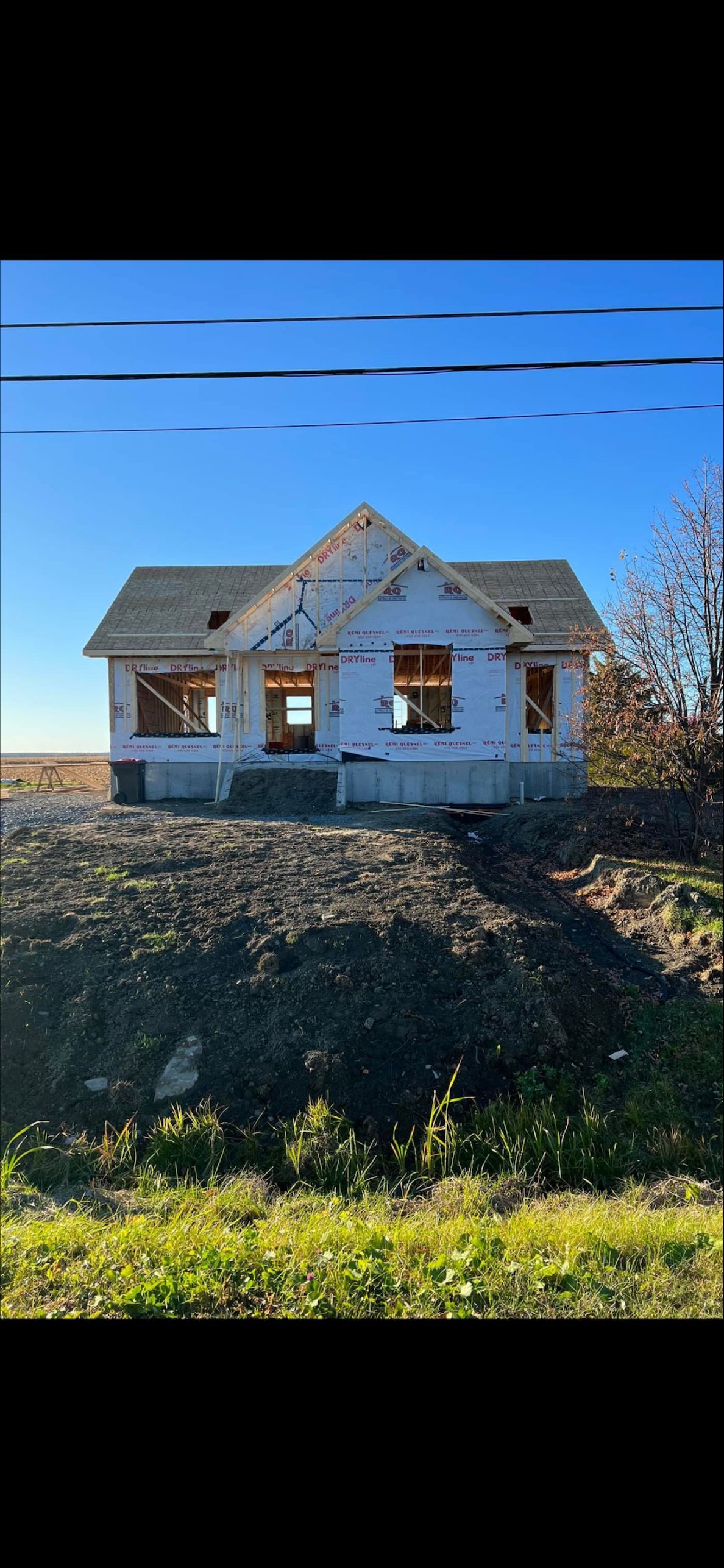 Sur une colline, sous un ciel dégagé, se dresse une structure de maison partiellement construite, avec un revêtement extérieur blanc et un toit de bardeaux.