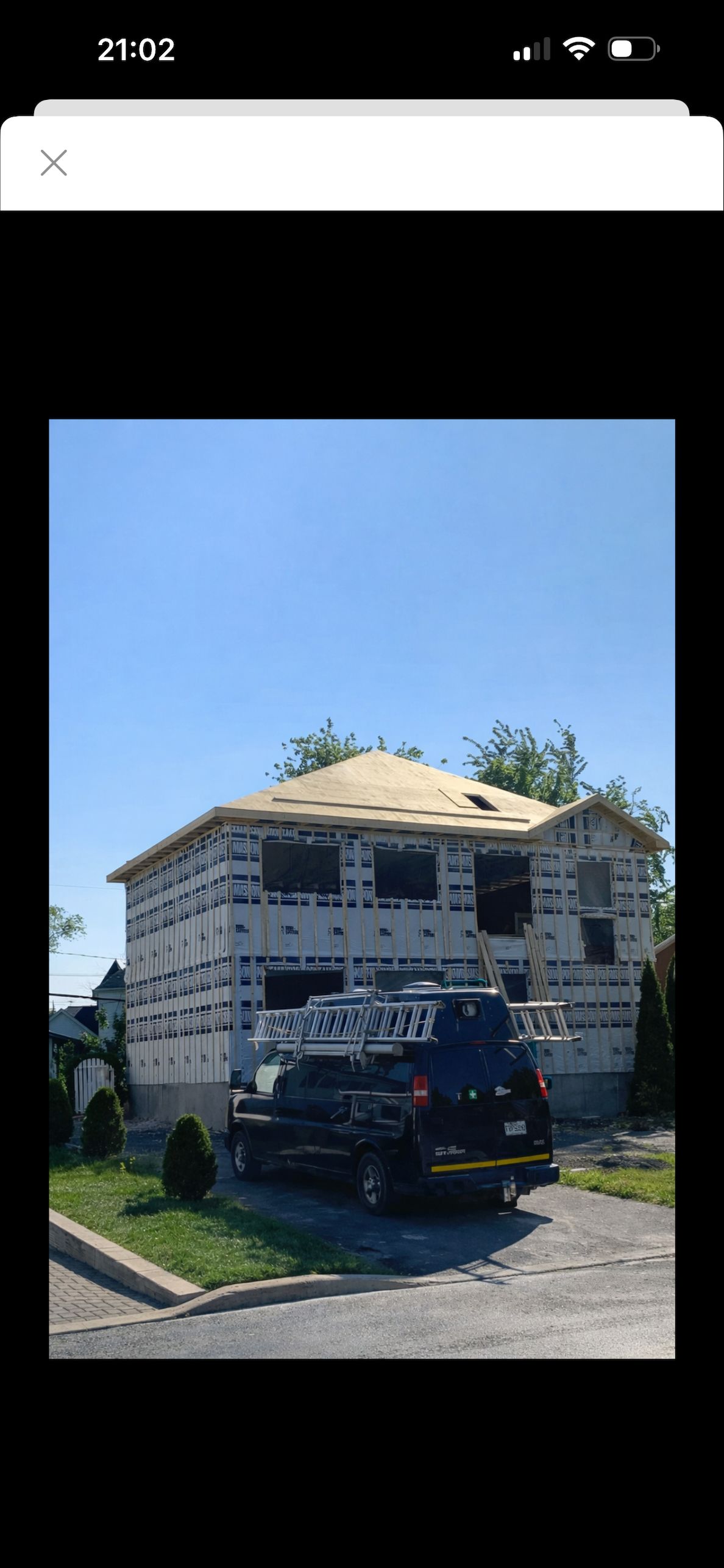 Une maison à deux étages en construction, avec des murs en parpaings blancs et un toit en contreplaqué récemment installé, un camion garé devant.