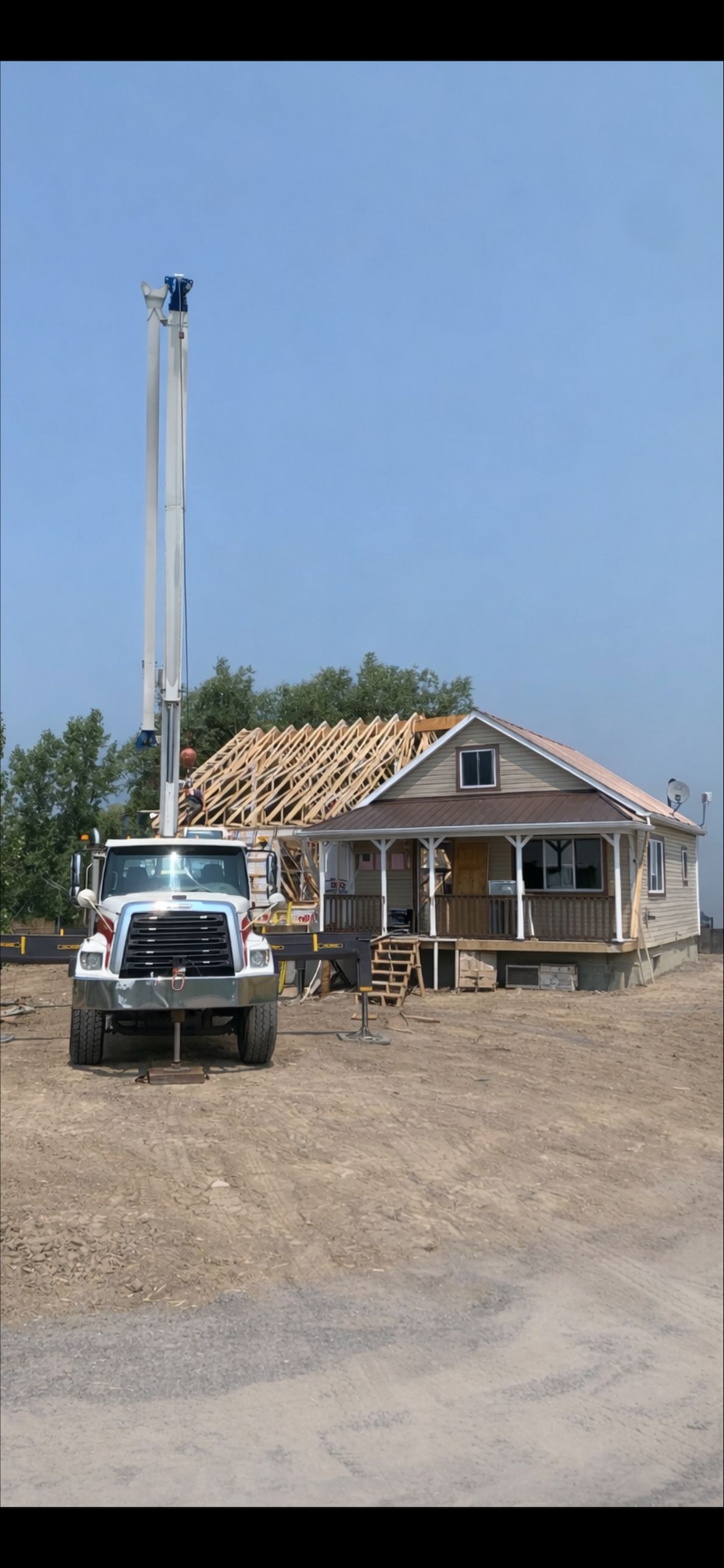 Un camion-pompe à béton blanc est stationné devant une petite maison en construction de toiture, sur un terrain vague.