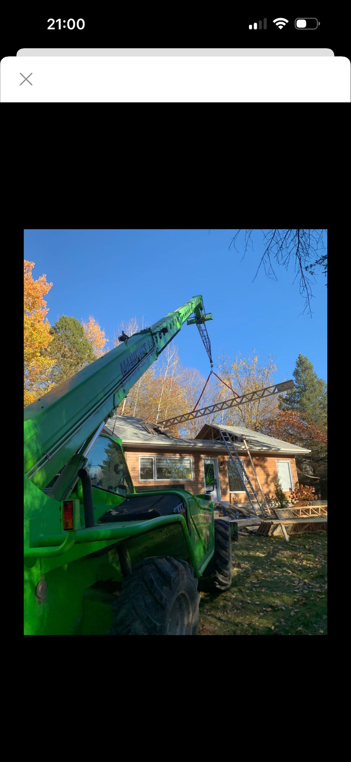Un chariot télescopique de chantier vert vif utilise un accessoire de grue pour soulever le toit d'un petit bâtiment en bois à l'extérieur.