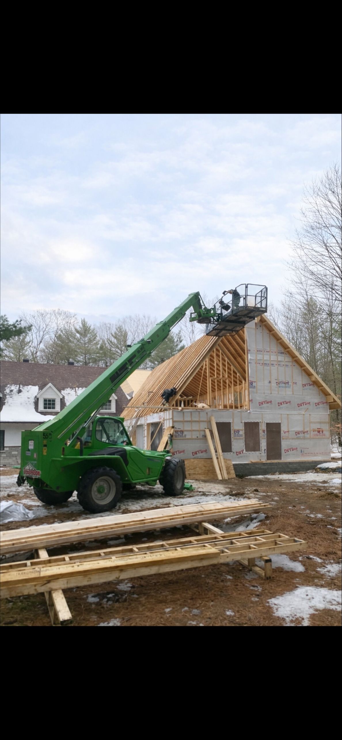Un chariot télescopique vert soulève un ouvrier installé sur une plateforme jusqu'au toit d'une maison à ossature bois en construction sur un terrain enneigé.