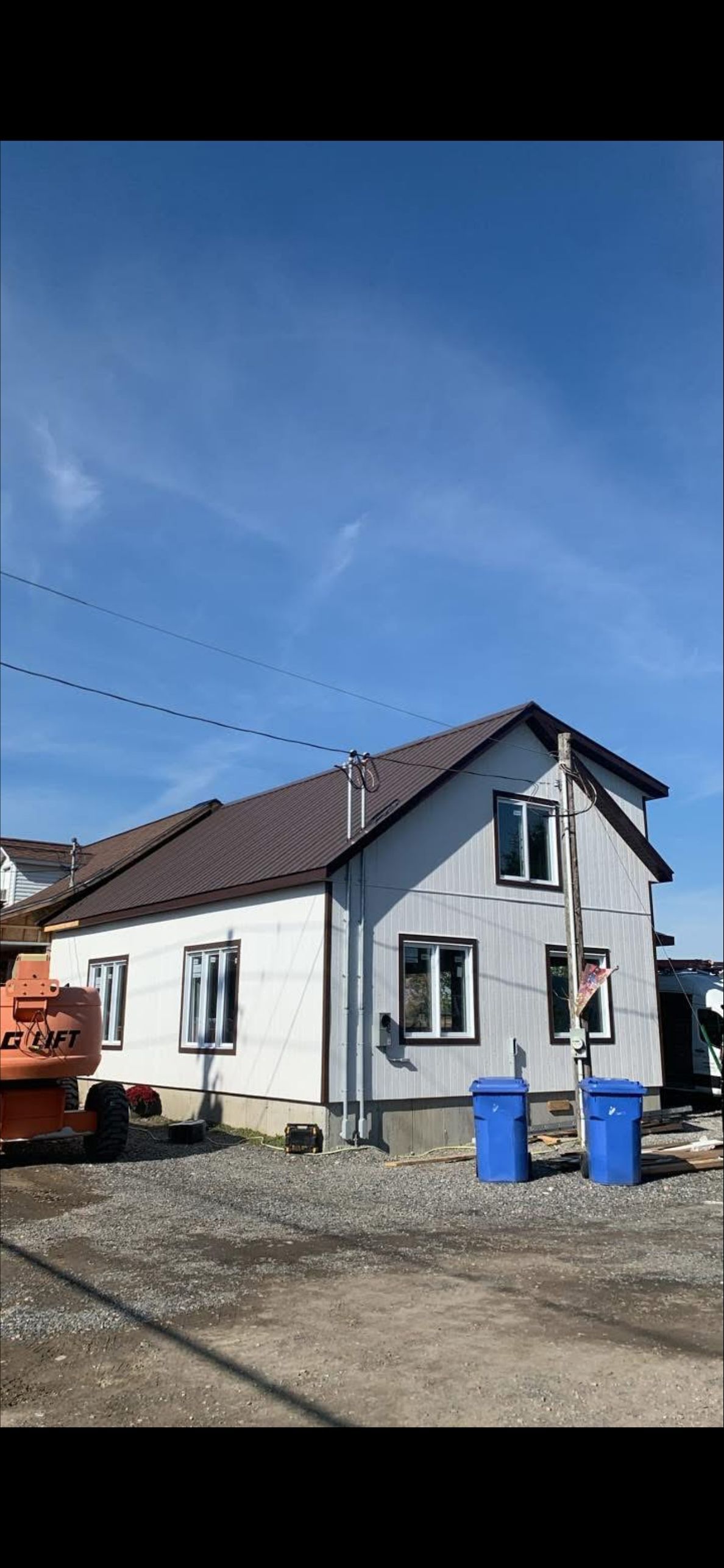 Une maison blanche sous un ciel bleu limpide, avec un toit marron, des fenêtres neuves et deux poubelles bleues devant.