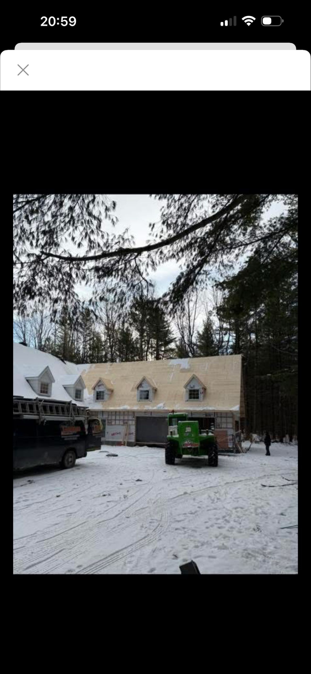 Des ouvriers du bâtiment travaillent sur le toit d'une grande maison dans un cadre boisé et enneigé, avec une camionnette et un tracteur à proximité.