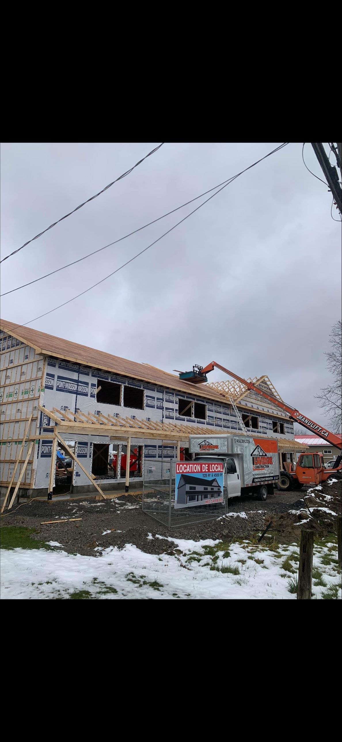 Des ouvriers du bâtiment utilisent une grue pour installer un toit sur un immeuble de deux étages sous un ciel nuageux.