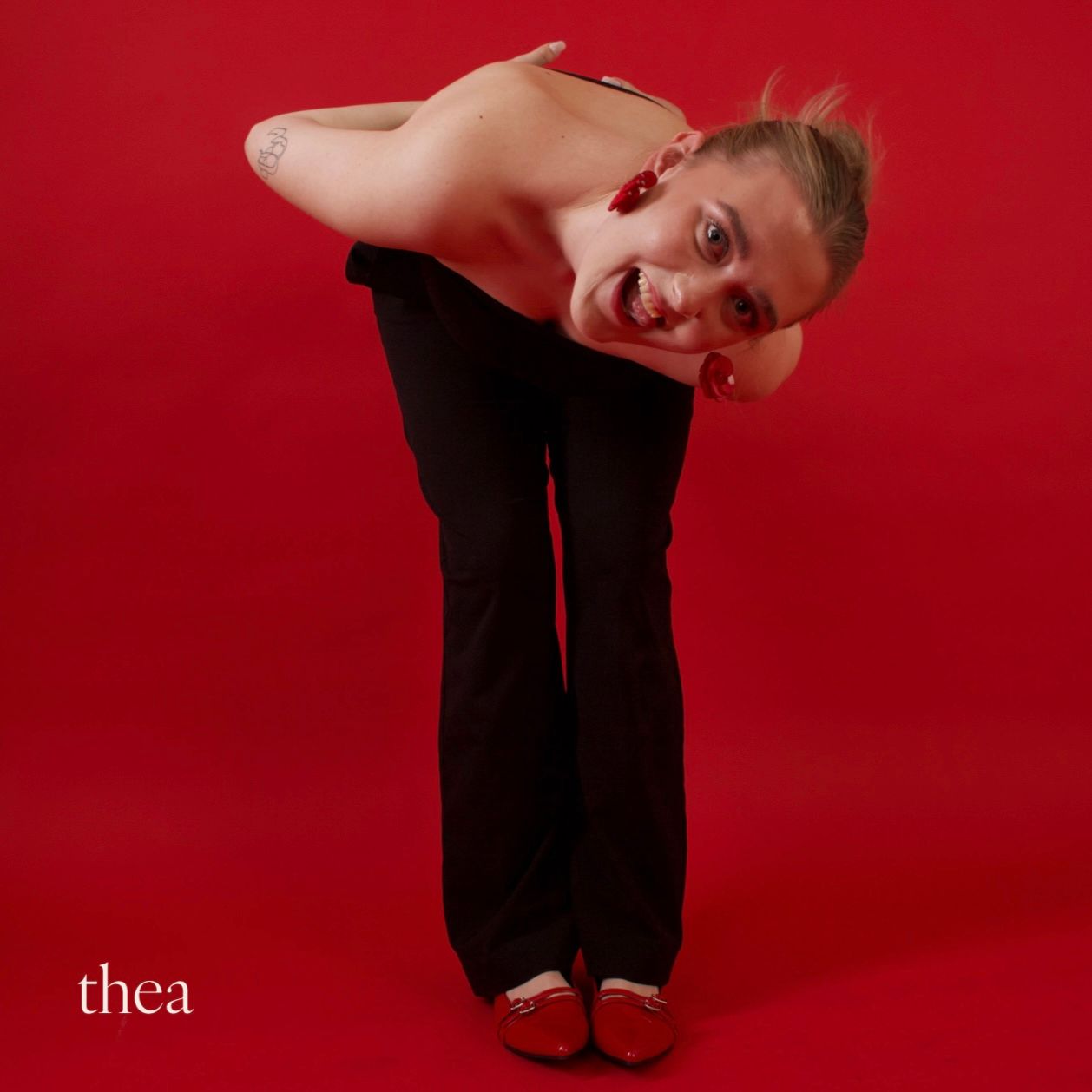 Person in black pants and red shoes, bends forward, arms crossed, mouth open, red background.