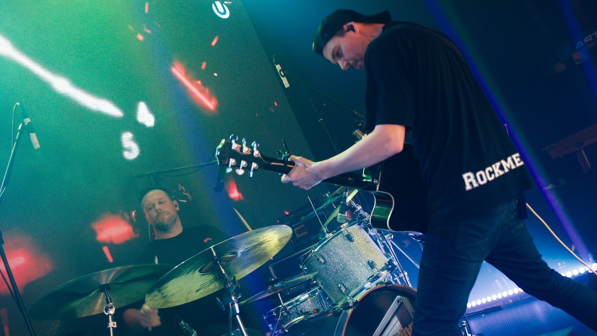 Guitarist and drummer performing on stage with colorful lighting. The guitarist wears a black shirt that says 