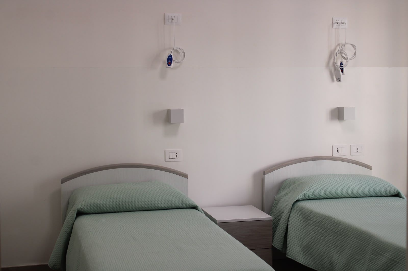 Due letti singoli con lenzuola verdi in una semplice stanza d’ospedale, separati da un comodino.