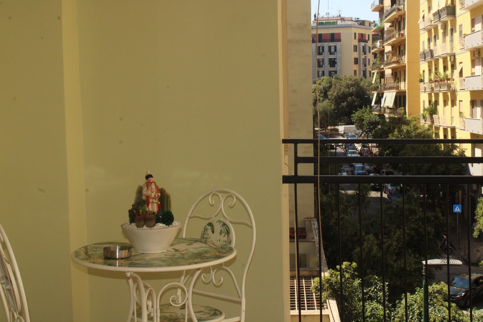 Balcone con un tavolino e una sedia in metallo bianco accanto a una parete gialla, affacciato sui palazzi vicini.
