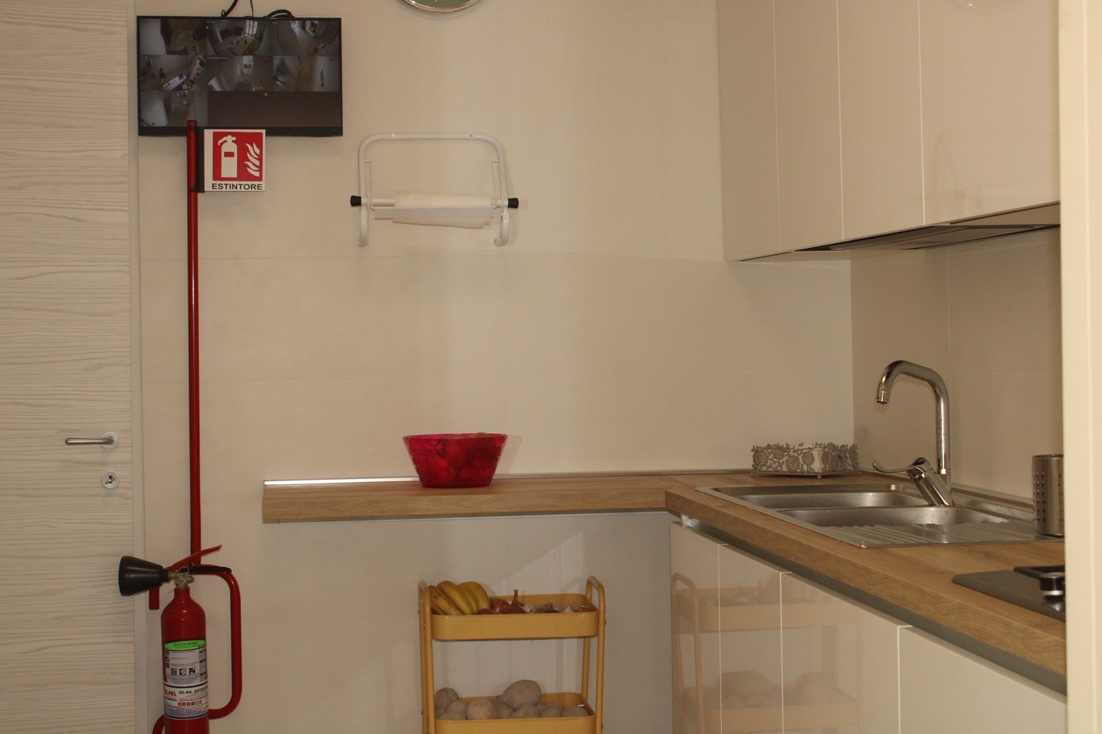 Piccola cucina con lavello, piano di lavoro, mobili, estintore e una ciotola rossa sul tavolo.