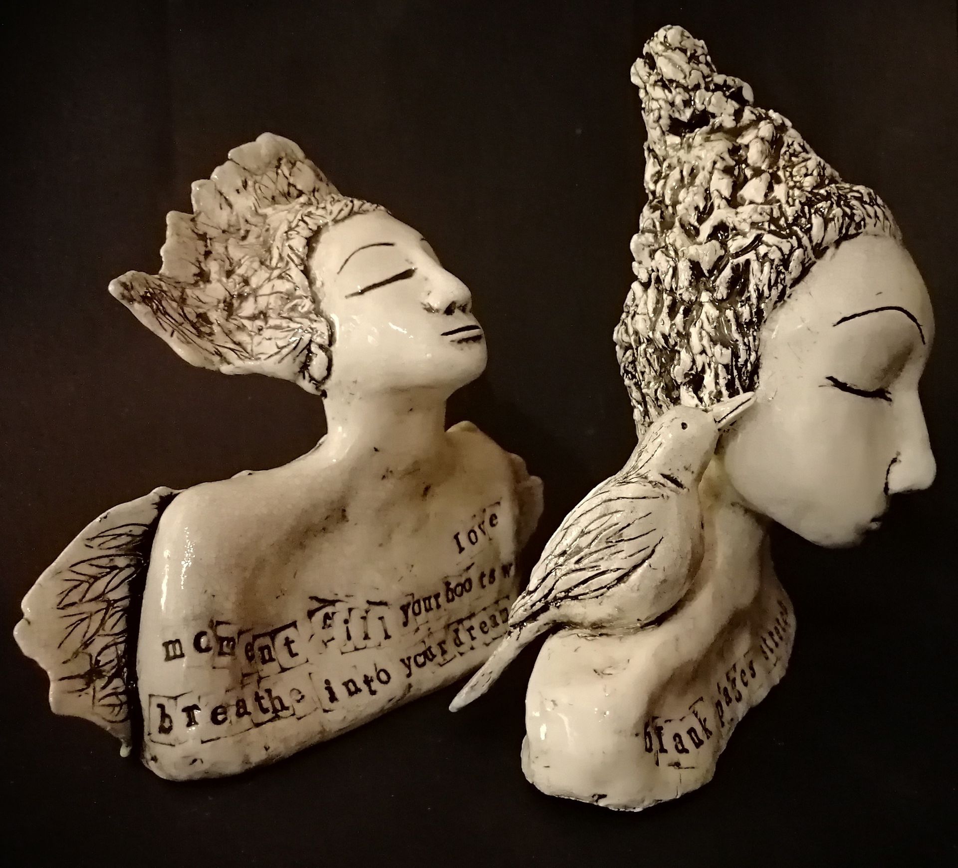 Two white ceramic busts of figures with closed eyes, bird perched on one, text inscriptions.