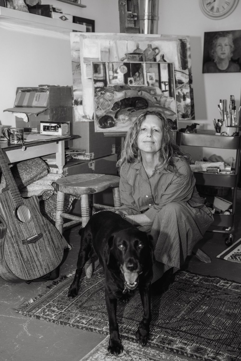 Woman and black dog in an art studio. A painting, guitar, and desk visible in a cluttered space.