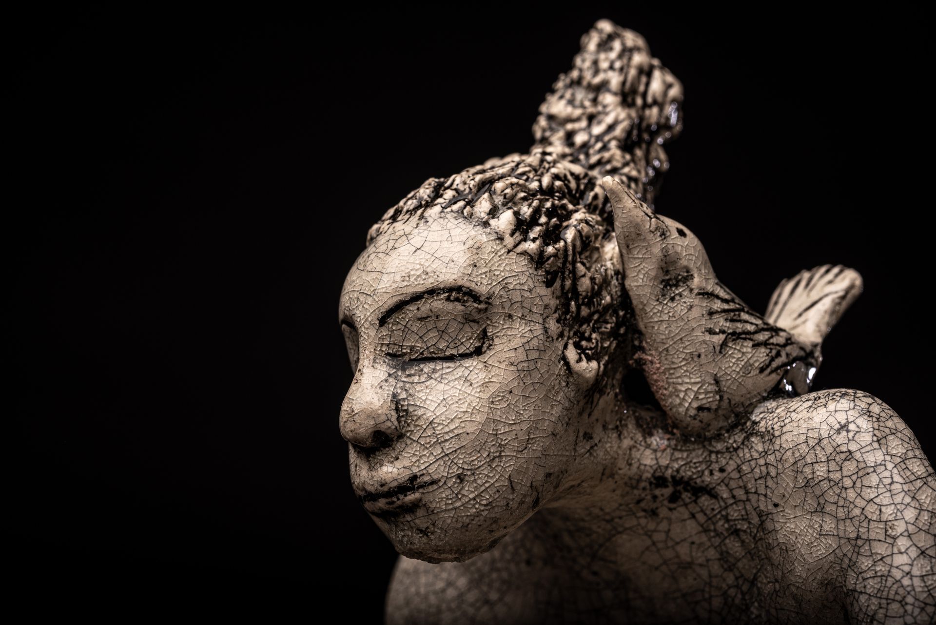 Sculpture of a woman with closed eyes, a bird perched on her shoulder, against a black background.