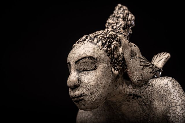 Stone-like bust of a woman with closed eyes, bird perched on her shoulder, against black background.