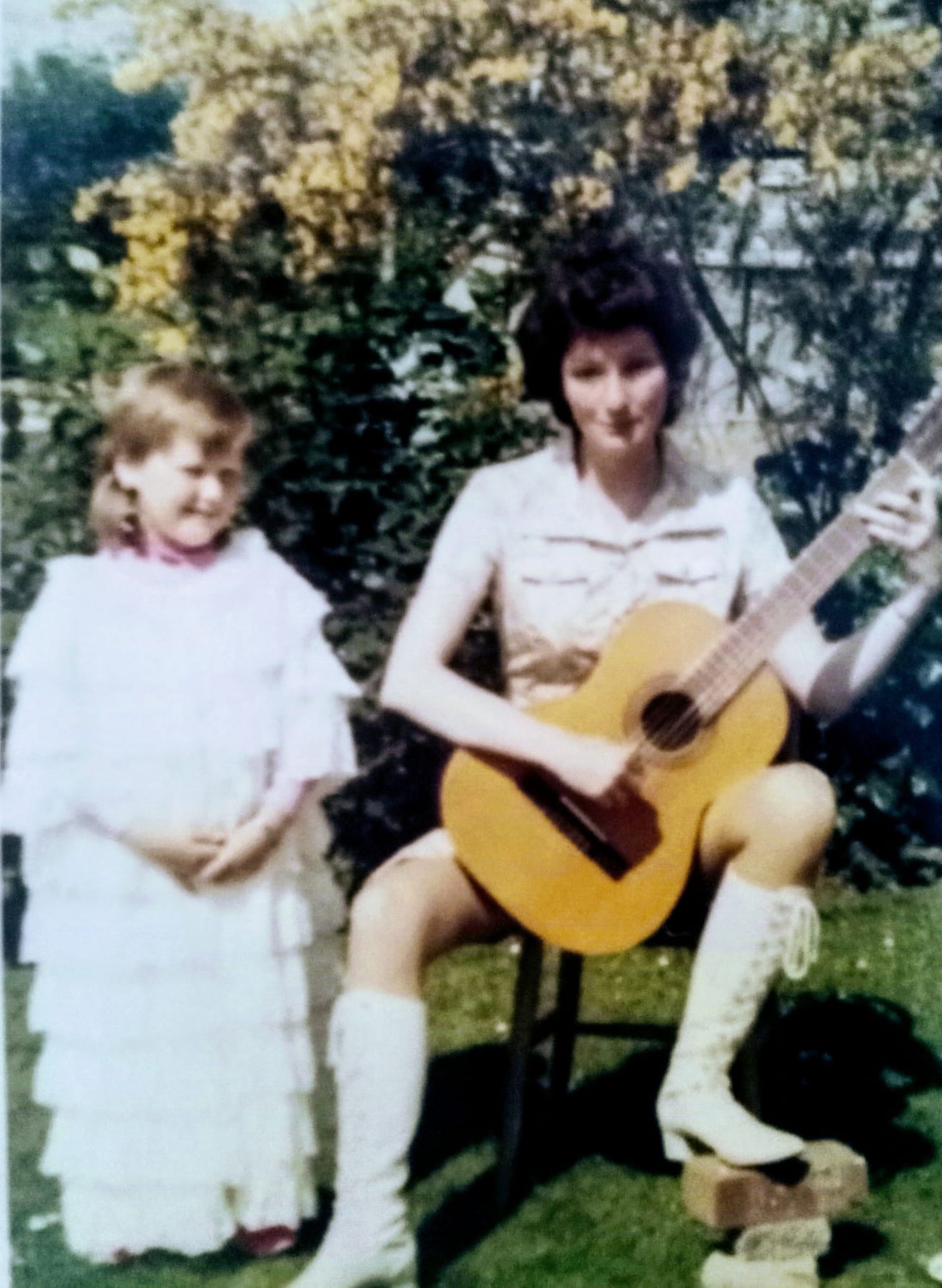 Woman playing guitar, seated outdoors, child in white dress beside her.