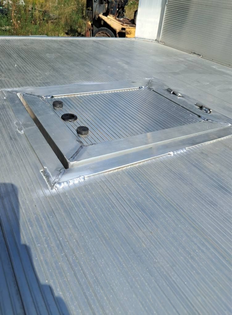Rooftop metal hatch with three vents on a corrugated silver roof, near a building wall.
