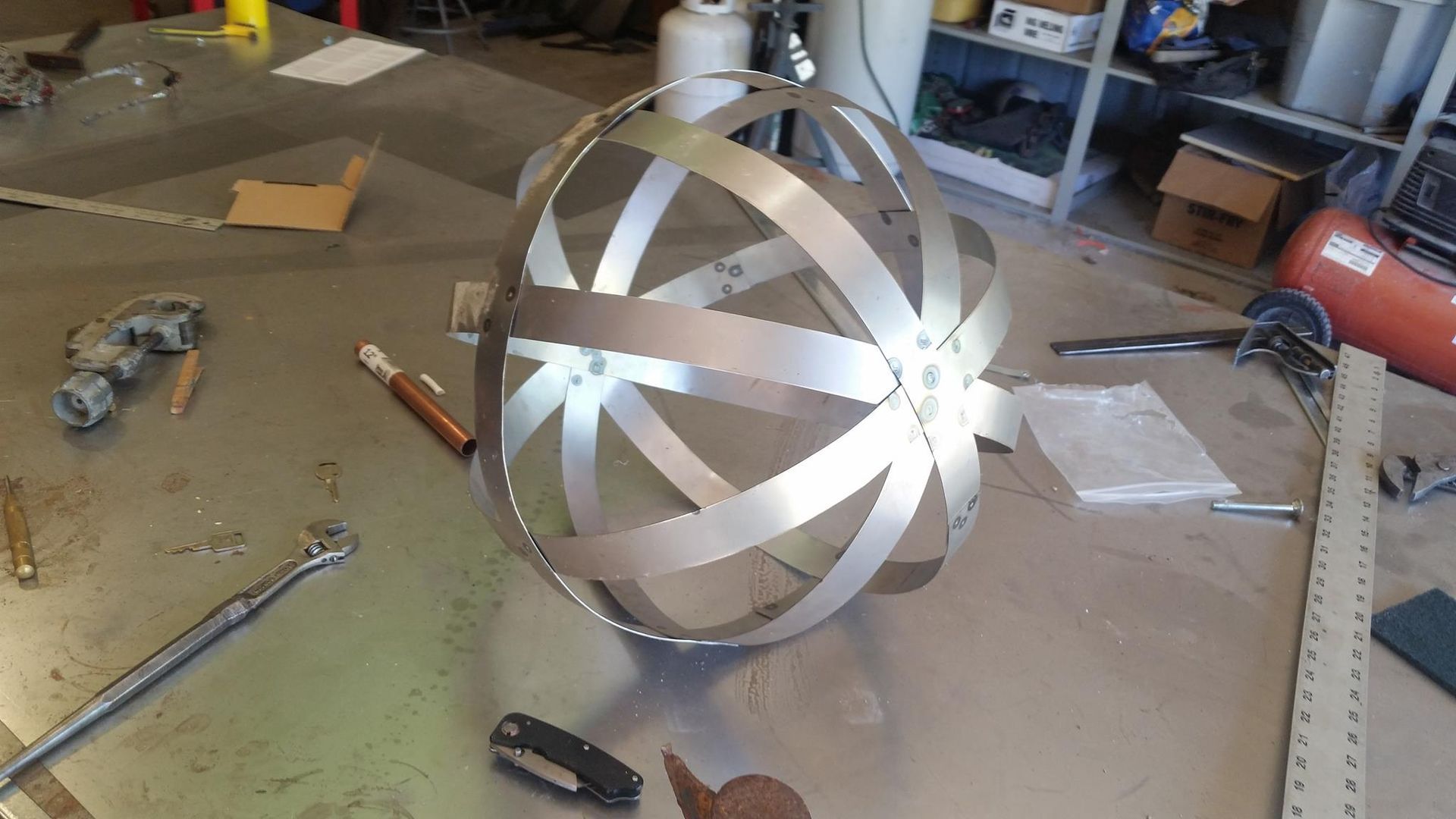 Metal spherical sculpture made of shiny strips on a workshop table, with tools and materials around it