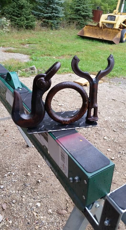 Rusty metal bracket with a circular ring mounted on a green beam outdoors on gravel, with trees and machinery in background
