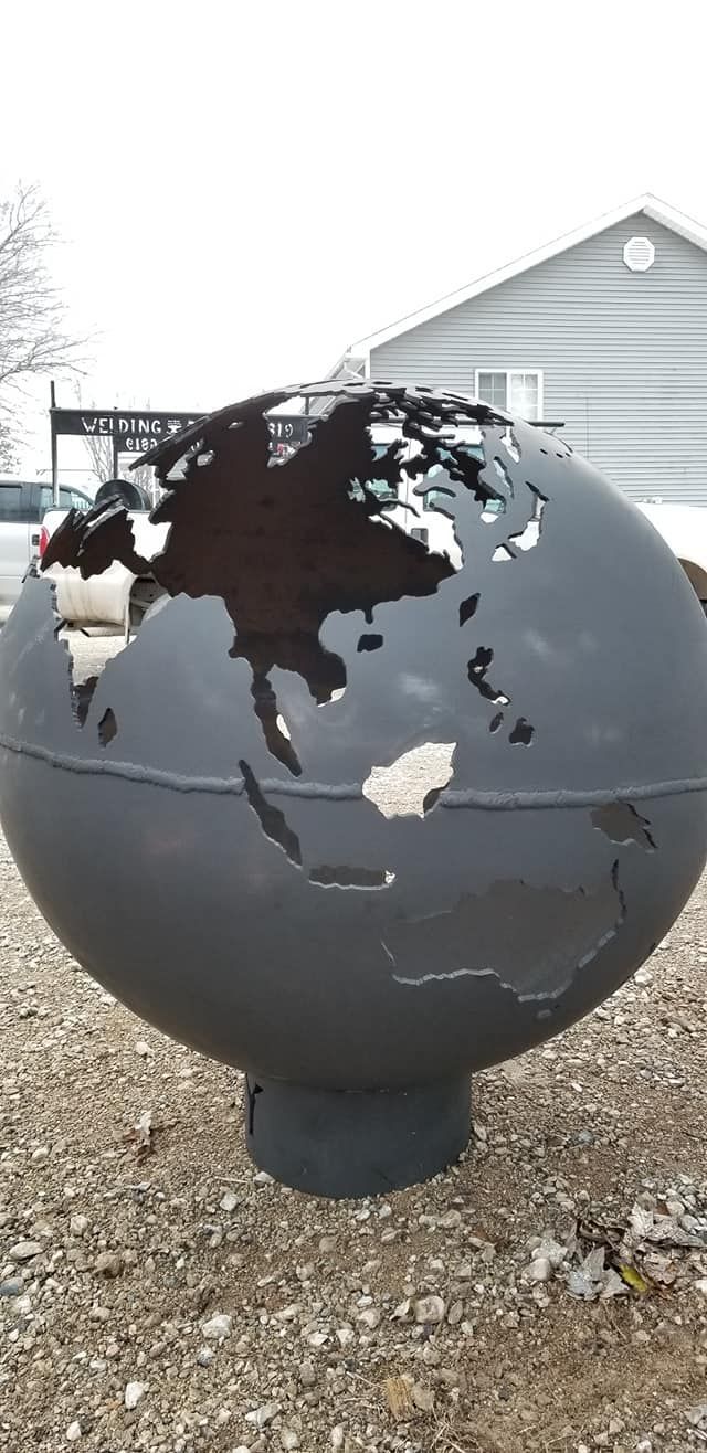 Black metal globe sculpture on a gravel lot in front of a light gray building.