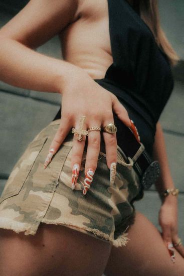 Close-up of a person in a black top and camo skirt, wearing multiple rings and long nails.