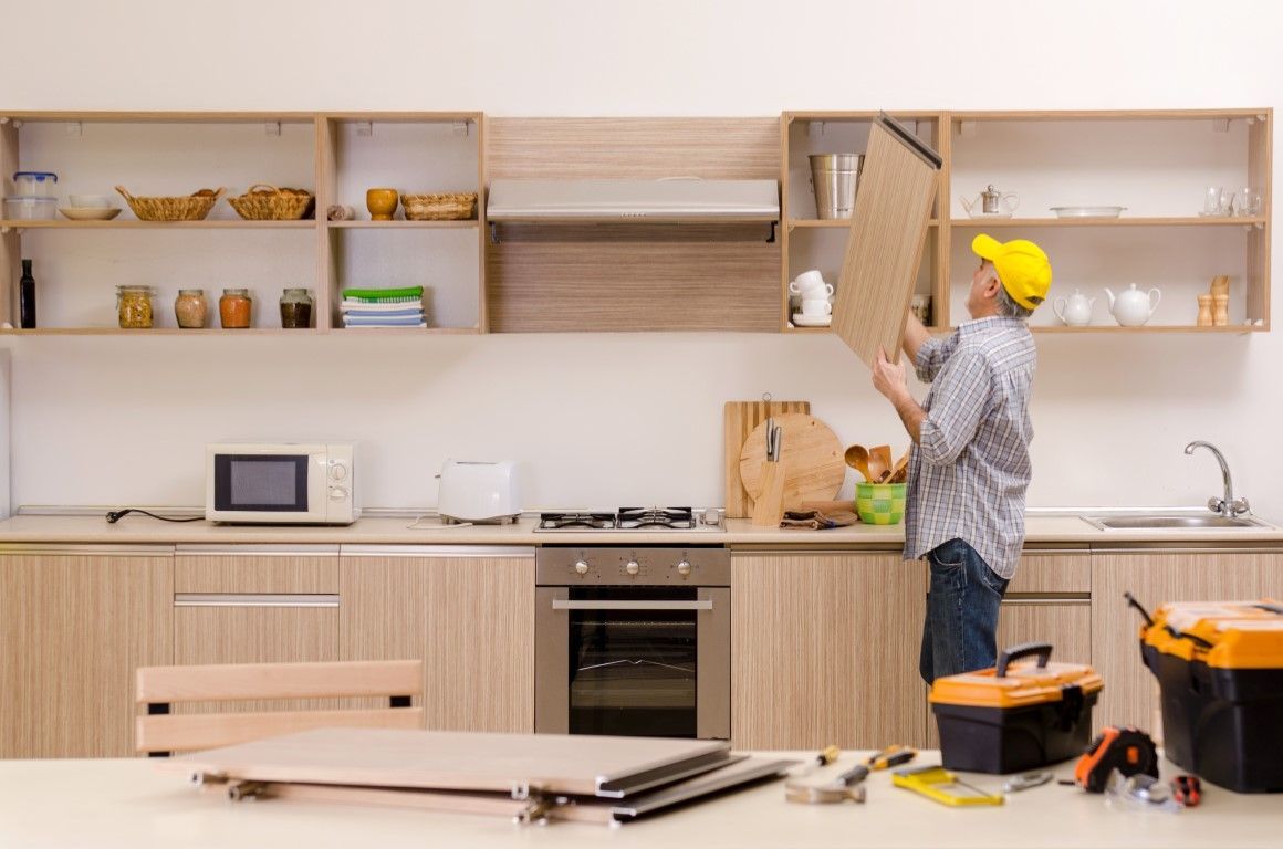 Person installing shelving in a bright kitchen with light wood cabinets and tools on the counter