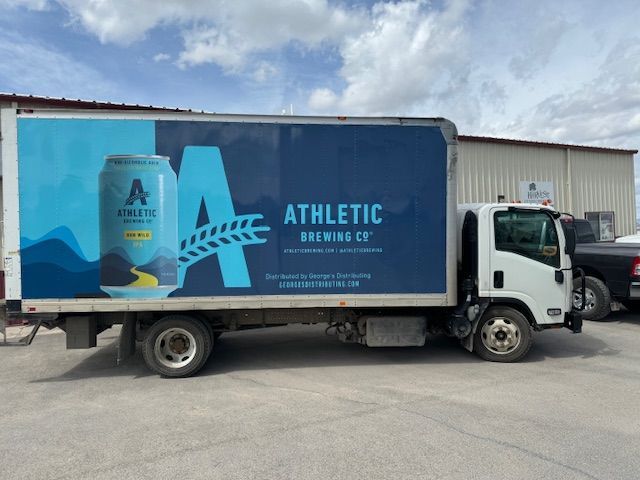 Delivery truck with Athletic Brewing Co. advertisement, featuring a blue beer can graphic and logo.