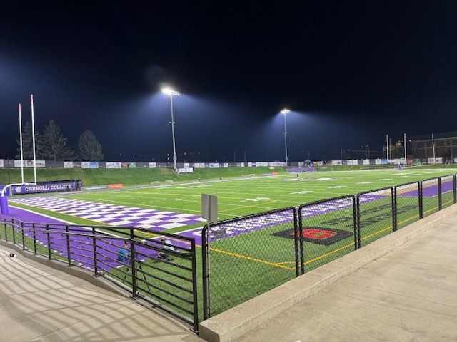 Football field at night, illuminated by stadium lights. Purple and white team colors visible.